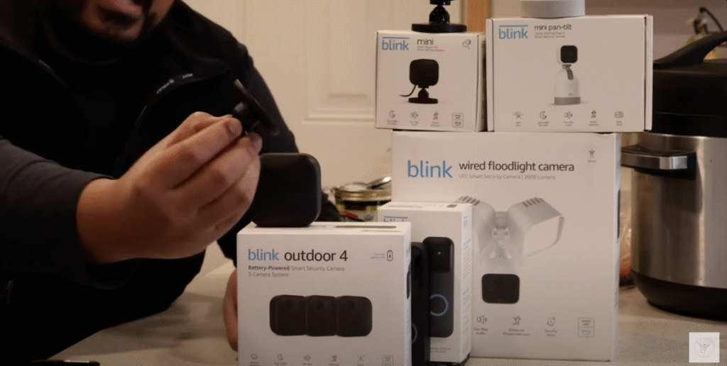 Person holding Blink camera with several Blink product boxes displayed, including Blink Outdoor 4, Mini, Mini Pan-Tilt, and Wired Floodlight.